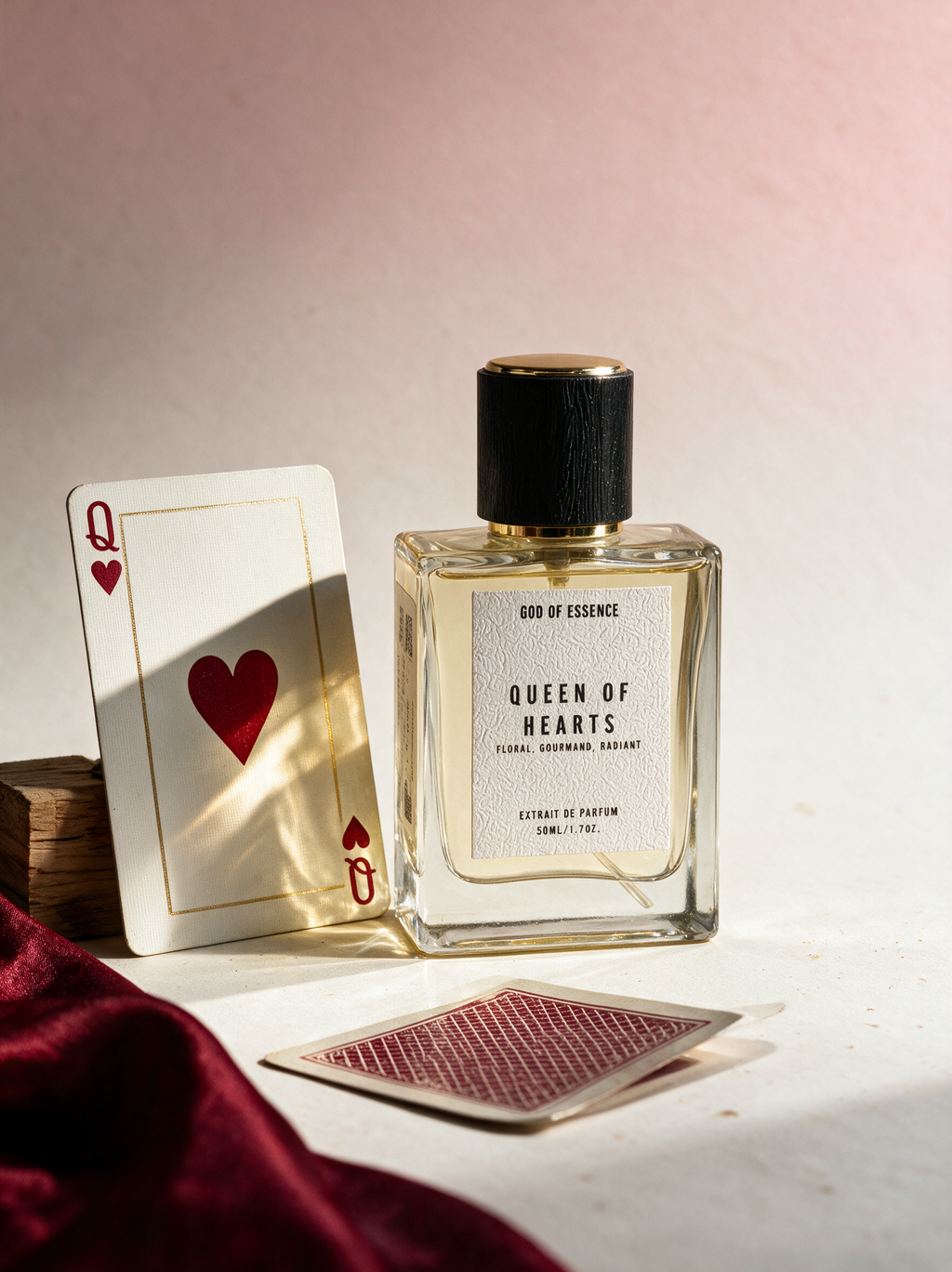 A clear glass perfume bottle labeled Queen of Hearts God of Essence, styled with a playing card, another card face down, and a draped burgundy fabric on a light surface against a soft pink background.